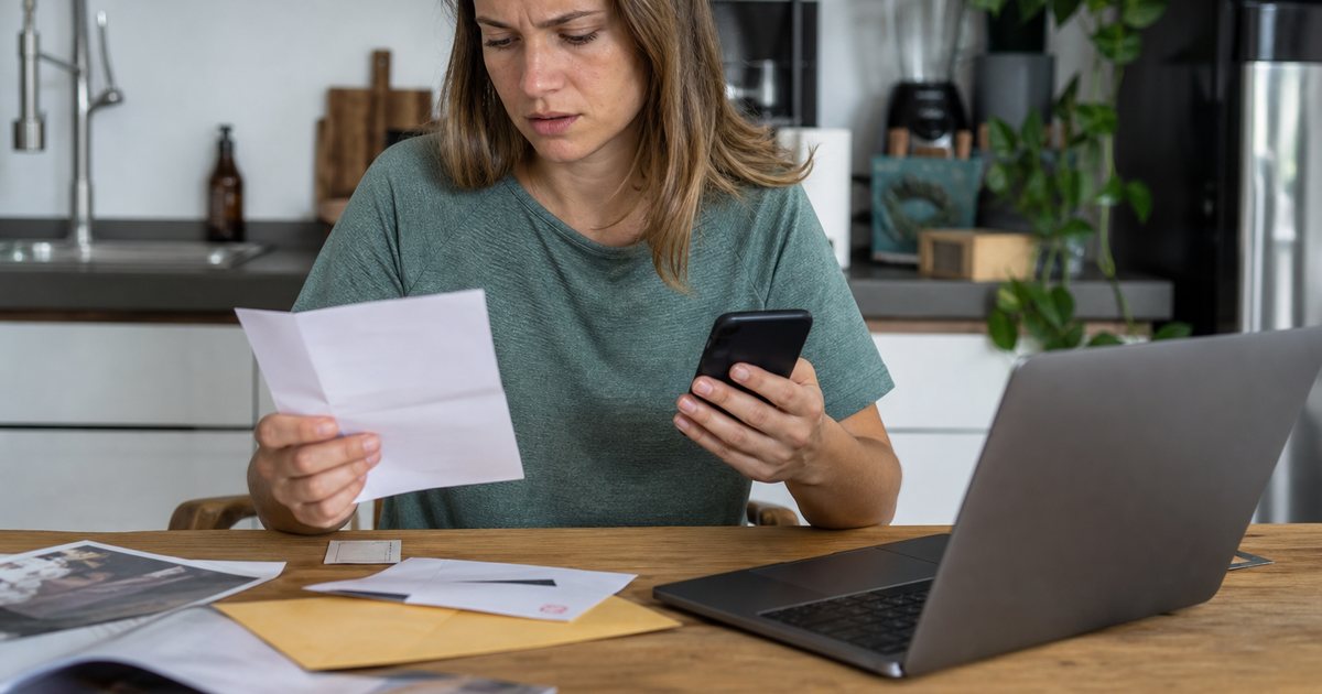 A freelance photographer at a kitchen table holding a phone to her ear, looking puzzled, blank envelopes and a closed laptop beside her, soft daylight