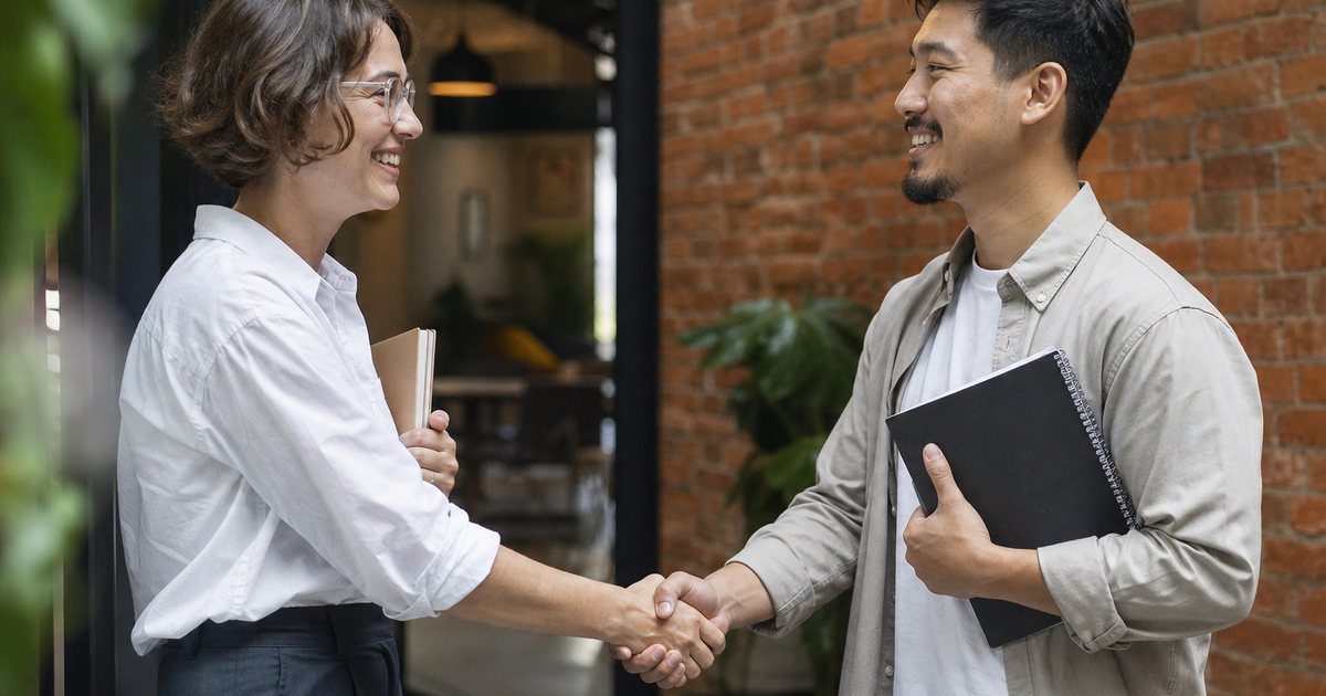 A freelance copywriter and a new client shaking hands at the entrance of a small co-working space, both smiling, notebooks under their arms, brick wall and plants
